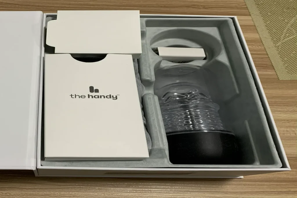 Geöffnete The Handy 2 Pro Verpackung mit Gerät, TrueGrip Cup und Zubehörteilen im Innenfach.