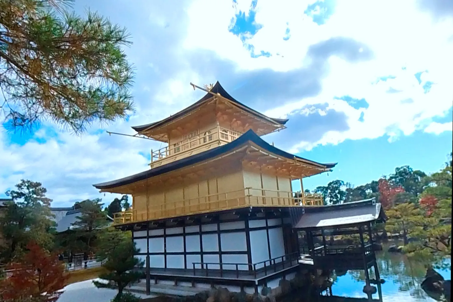 The Golden Pavilion of Kinkaku-ji in Kyoto reflected in a calm pond under a clear blue sky, surrounded by pine trees and autumn foliage.