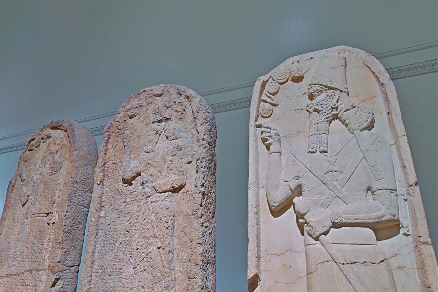 Assyrian stone relief displayed in the British Museum, showing a bearded king holding a staff on the right and a weathered warrior figure on the left, conveying the weight of ancient civilization and time.