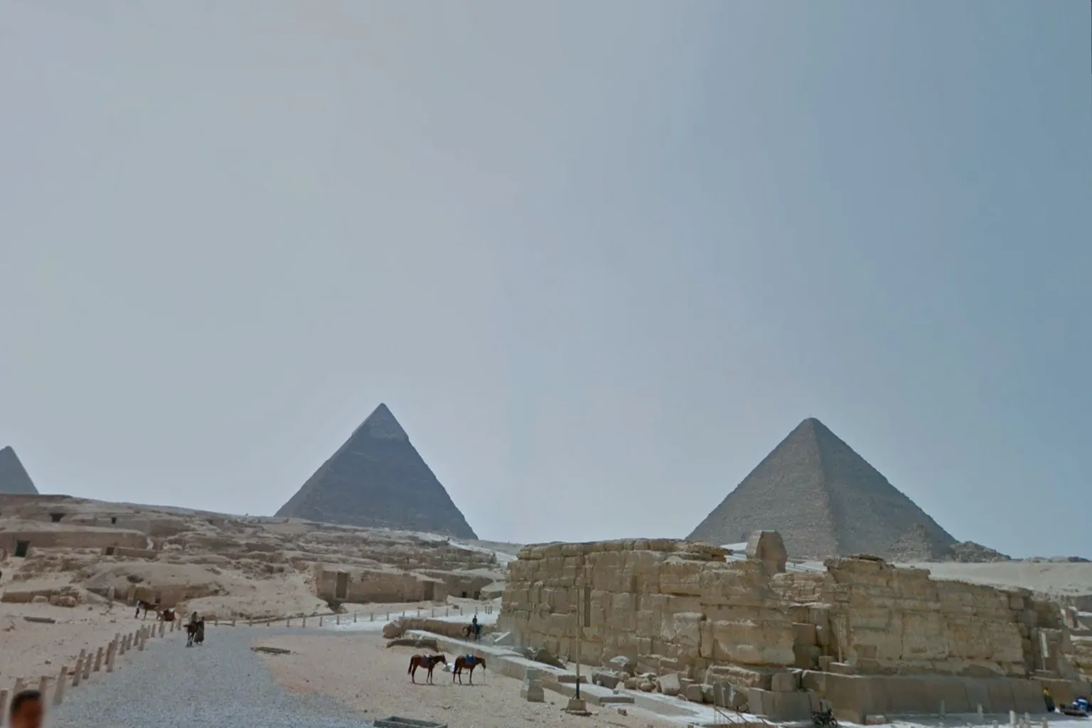 The Pyramids of Giza in Egypt, towering ancient stone structures rising from sandy dunes, with sunlight illuminating the scene and camels and tourists visible in the distance.