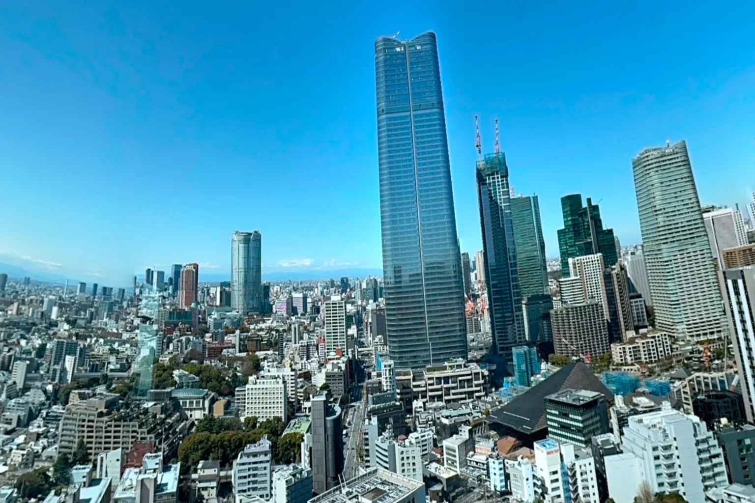 Daytime cityscape of Tokyo viewed from Tokyo Tower, with dense skyscrapers stretching toward the horizon and distant mountains visible under a clear sky.