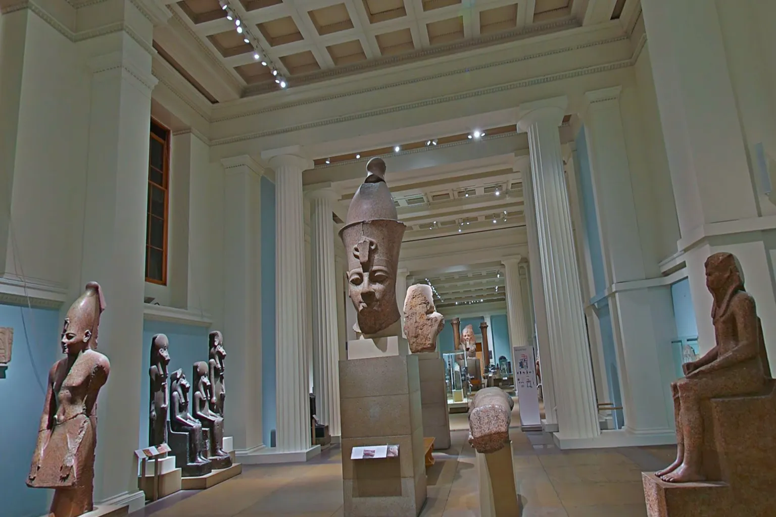 Interior of the British Museum in London, displaying ancient Egyptian statues and pharaoh busts as light filters through white colonnades.