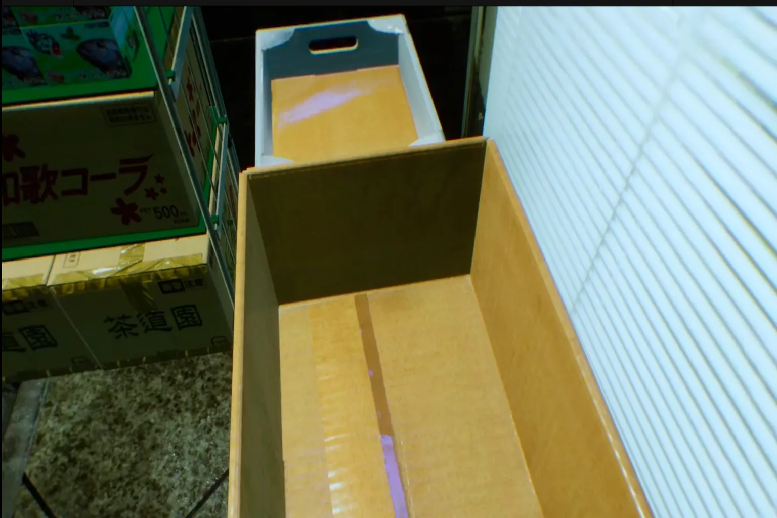 VRゲーム内でプレイヤーが空の段ボール箱に立ち、これから搬送任務に入ろうとしている。 周囲には日本語の表示がある貨物箱が積み上げられ、シーンの細部までリアルに描写されている。