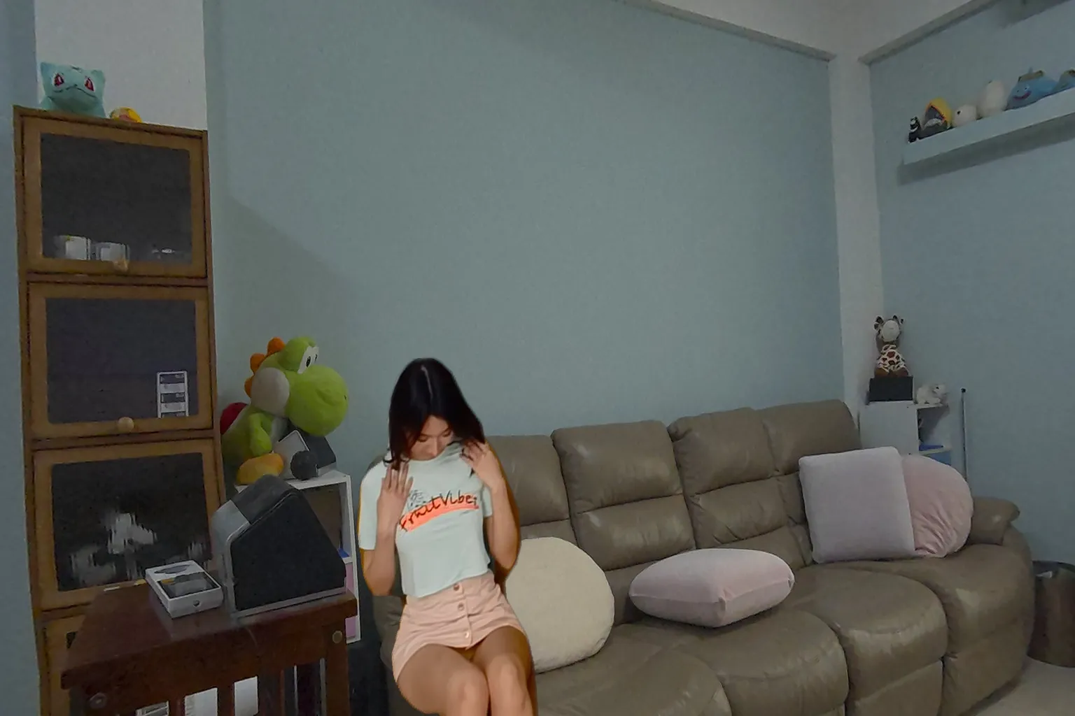 亞洲女孩穿著淺綠色短T與粉紅短裙,坐在沙發上低頭拉衣服,背景有玩偶與抱枕